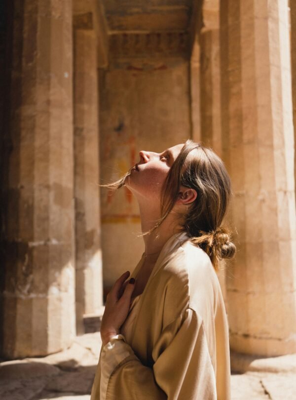 A woman in a beige coat looks up inside ancient Egyptian temple ruins.